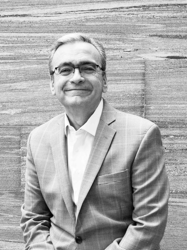 Smiling man in glasses and checked blazer against a textured wall.