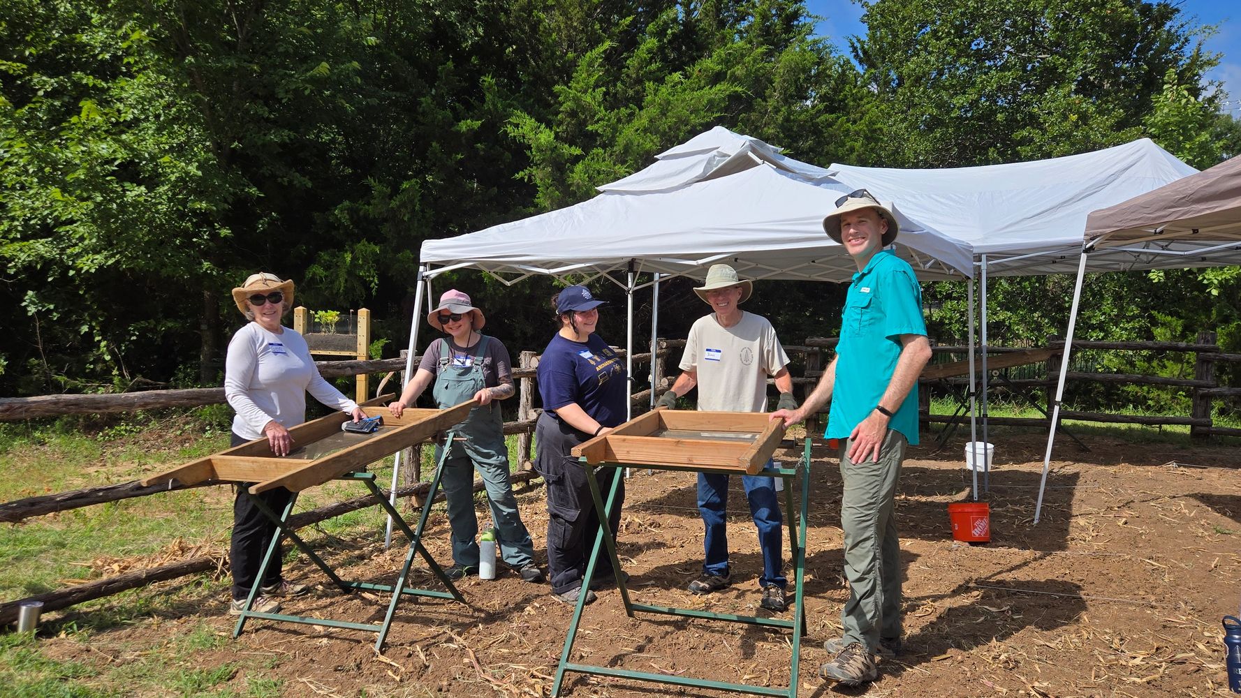 A group of people in outdoor clothing stand next to screens used for sifting dirt.