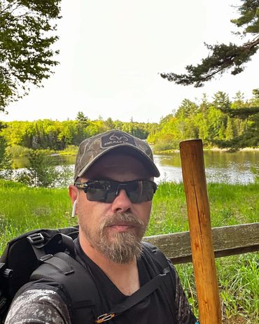 Man with sunglasses and cap holding wooden stick near a lake surrounded by greenery.
