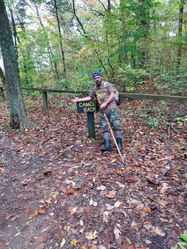Man in hiking gear pointing at a sign that says Camel's Back in a forest.