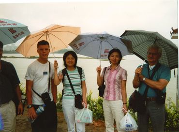 Four people standing outdoors holding umbrellas near a water body.
