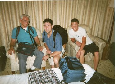 Three men with backpacks sitting in a room with luggage around.