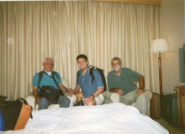 Three men sitting on chairs in a hotel room with backpacks and luggage.