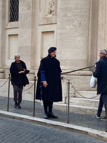 Man in historical costume stands on a cobblestone street near two pedestrians.