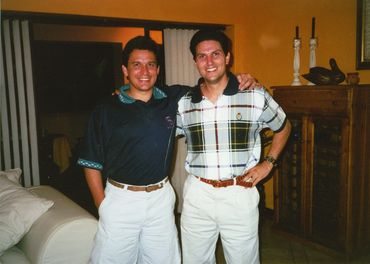 Two men smiling indoors, dressed in casual polo shirts and white pants.