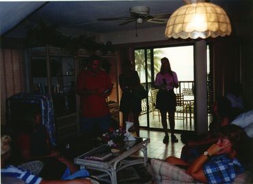 People gathered in a dimly lit living room with a balcony view of palm trees.