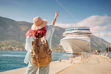 Traveler with red hair and backpack making peace sign at a dock with a cruise ship.