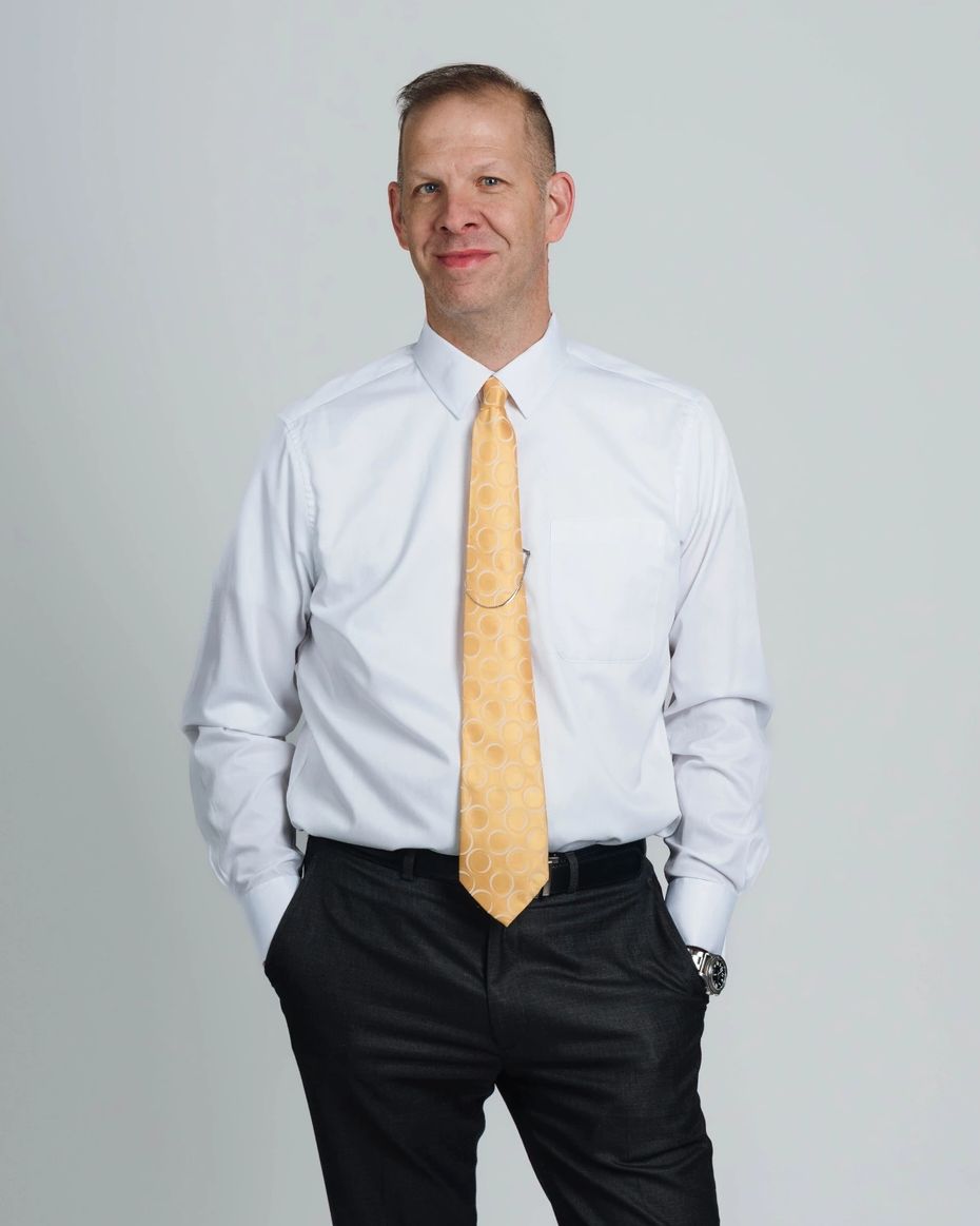 A man in formal attire stands confidently with hands in pockets against a plain background.