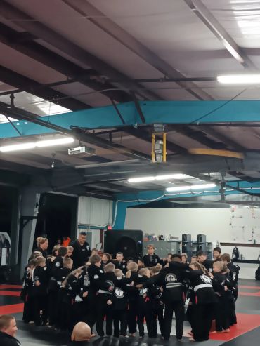 Children in martial arts uniforms gather in a circle inside a training gym.