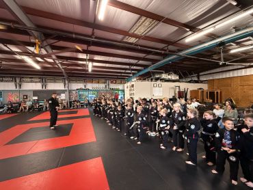 Children in martial arts uniforms line up in a dojo with an instructor addressing them.
