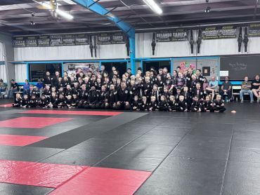Large group of martial artists posing in a gym with championship belts on the wall.