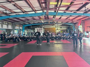 Martial arts class with instructors addressing a large group of students sitting on mats.