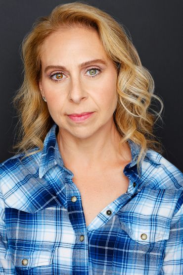 Woman with curly blonde hair wearing a blue plaid shirt against a dark background.