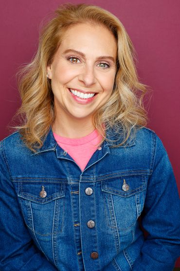 Smiling woman with blonde hair wearing a denim jacket and pink shirt against a purple background.
