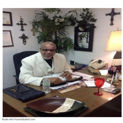Man in white suit sitting at desk with religious crosses on the wall.