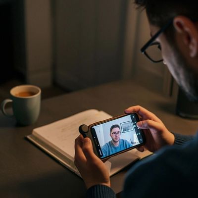 A man video chatting on his smartphone at a desk with a notebook and coffee.
