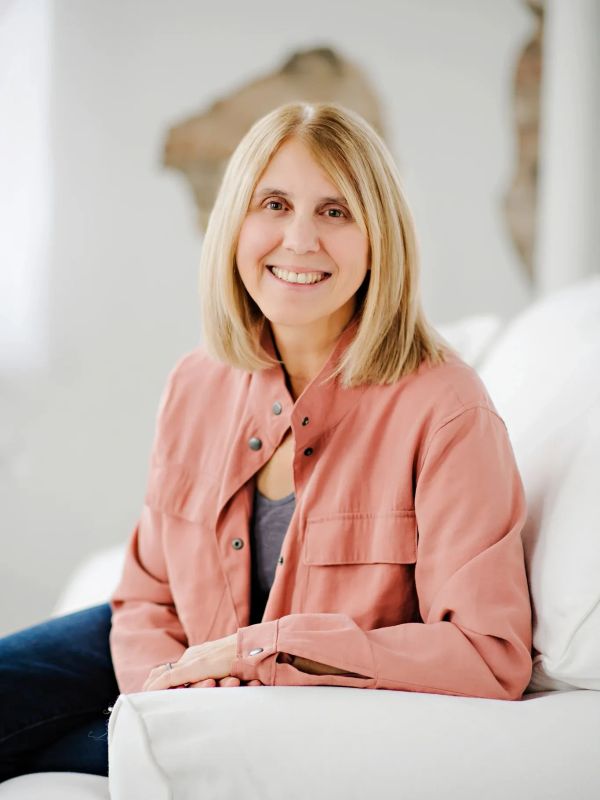 Smiling woman in a coral jacket sitting on a white couch.