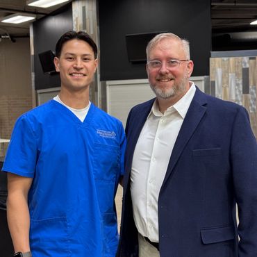 Two smiling men posing indoors, one in blue nursing scrubs, the other in a suit jacket.