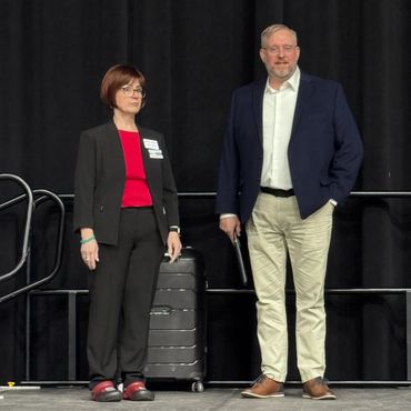 Two professionally dressed individuals standing on stage with a black suitcase behind them.