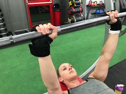 Woman lifting a barbell at the gym, wearing wrist support gloves.