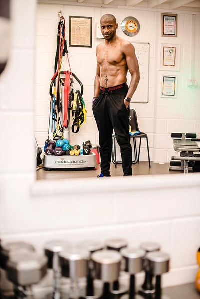 A shirtless man stands confidently in a gym filled with fitness equipment.
