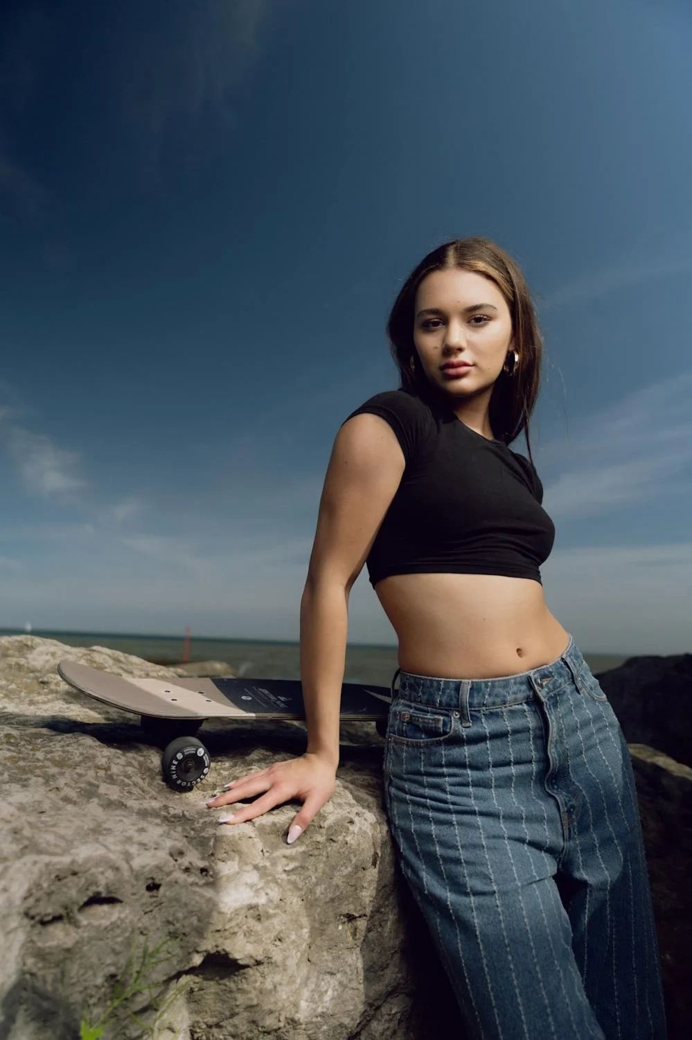 Young woman posing with skateboard on rocks under a clear sky.