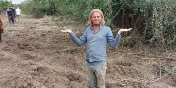 Mel standing in a deep hole in a field with trees around, shrugging his shoulders.