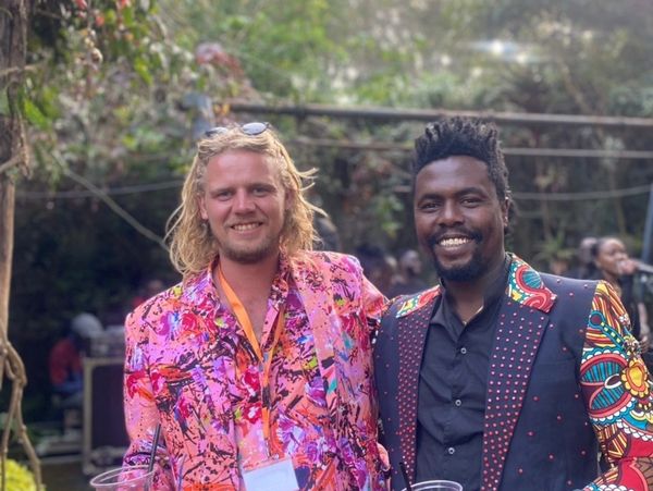 Two men smiling, dressed in colorful patterned suits, holding drinks outdoors.