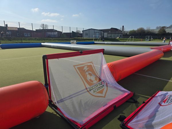 Inflatable soccer goals and barriers set up on an outdoor sports court under clear blue sky.