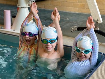 Three children in swim caps and goggles raising their arms in a pool.