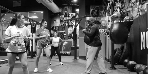 A boxing coach trains three women in a gym with punching bags.