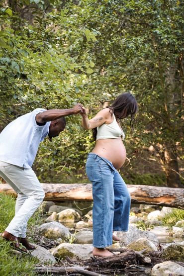 Pregnant woman crossing rocks with partner's help in a natural setting.