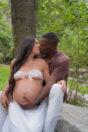 A loving couple shares a kiss, embracing the woman's pregnant belly in a serene outdoor setting.