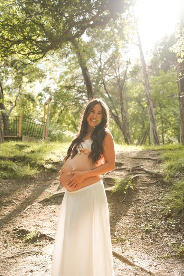 Pregnant woman smiling outdoors in sunlight, wearing a white skirt and floral top.
