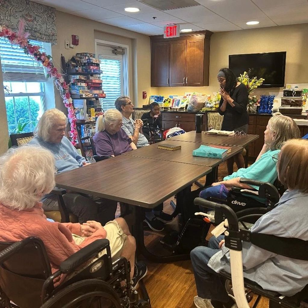 Group of elderly people gathered around a table listening to a woman.