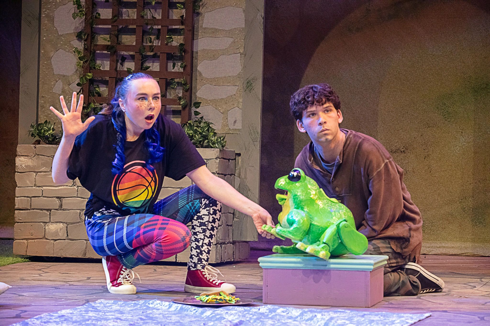 Two actors interact with a puppet frog on stage.