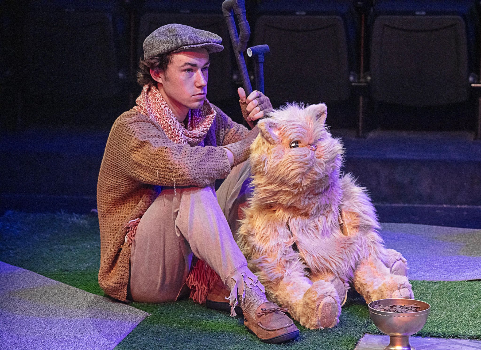 A young man in rustic clothes sitting on stage with a large furry cat puppet.