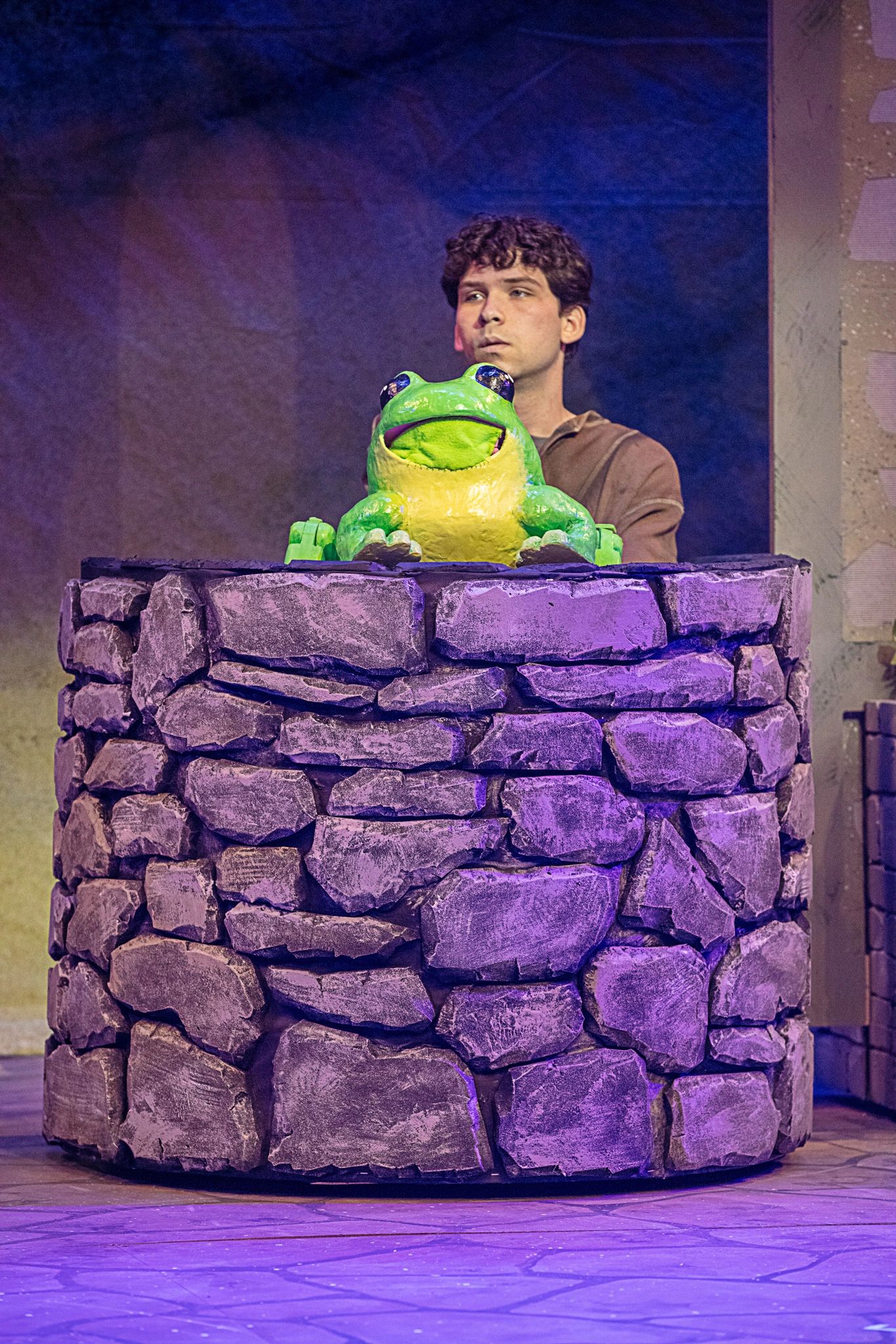 Man with a large frog puppet behind a stone well.