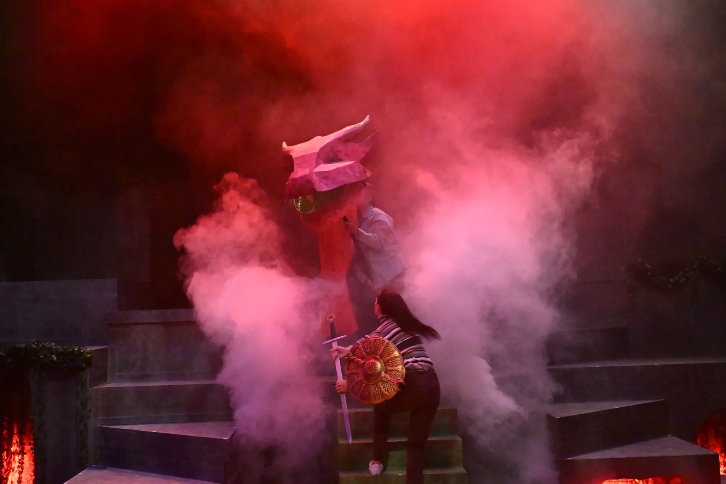 Person with sword and shield approaches another with a large mask amid smoke.