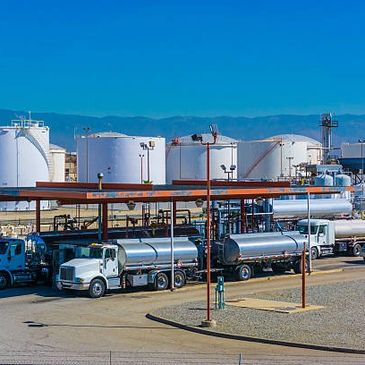 Fuel tanker trucks parked at an oil refinery with storage tanks in the background.