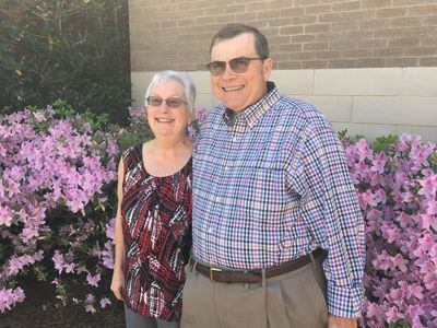 An elderly couple smiling in front of pink flowers outside a brick building.
