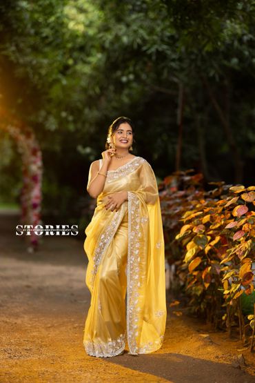 Woman in a yellow saree smiling outdoors among greenery and autumn leaves.