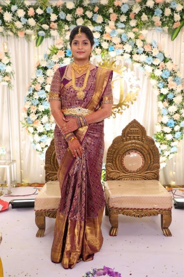 A woman in a traditional purple and gold saree stands before floral decorations.
