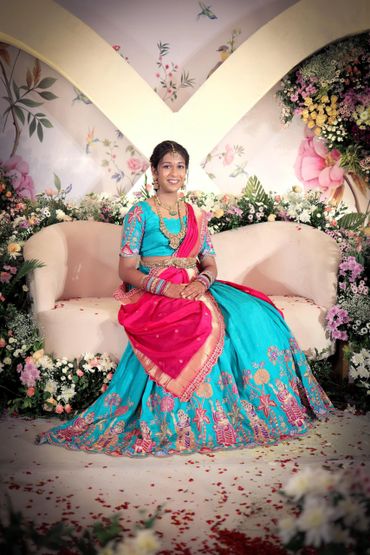 A woman in traditional Indian attire sits on a floral-decorated couch, smiling gracefully.