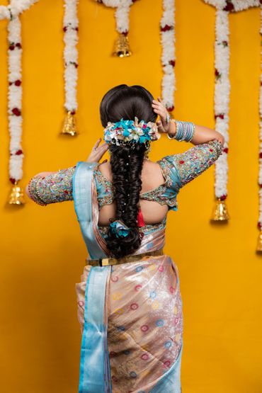 Woman in traditional saree with floral hair decoration against yellow backdrop.