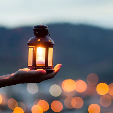 A hand holds a lantern, with light reflecting across a landscape at dusk.