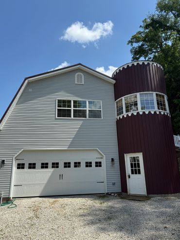 A modern barn-style house with a white garage and a tall maroon silo under a blue sky.