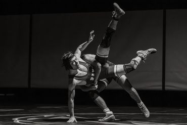 Two wrestlers compete intensely on the mat in a dramatic black and white scene.