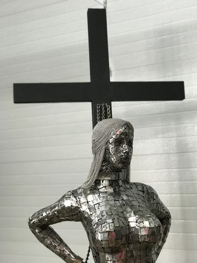 Metallic sculpture of a woman with a black cross in the background.