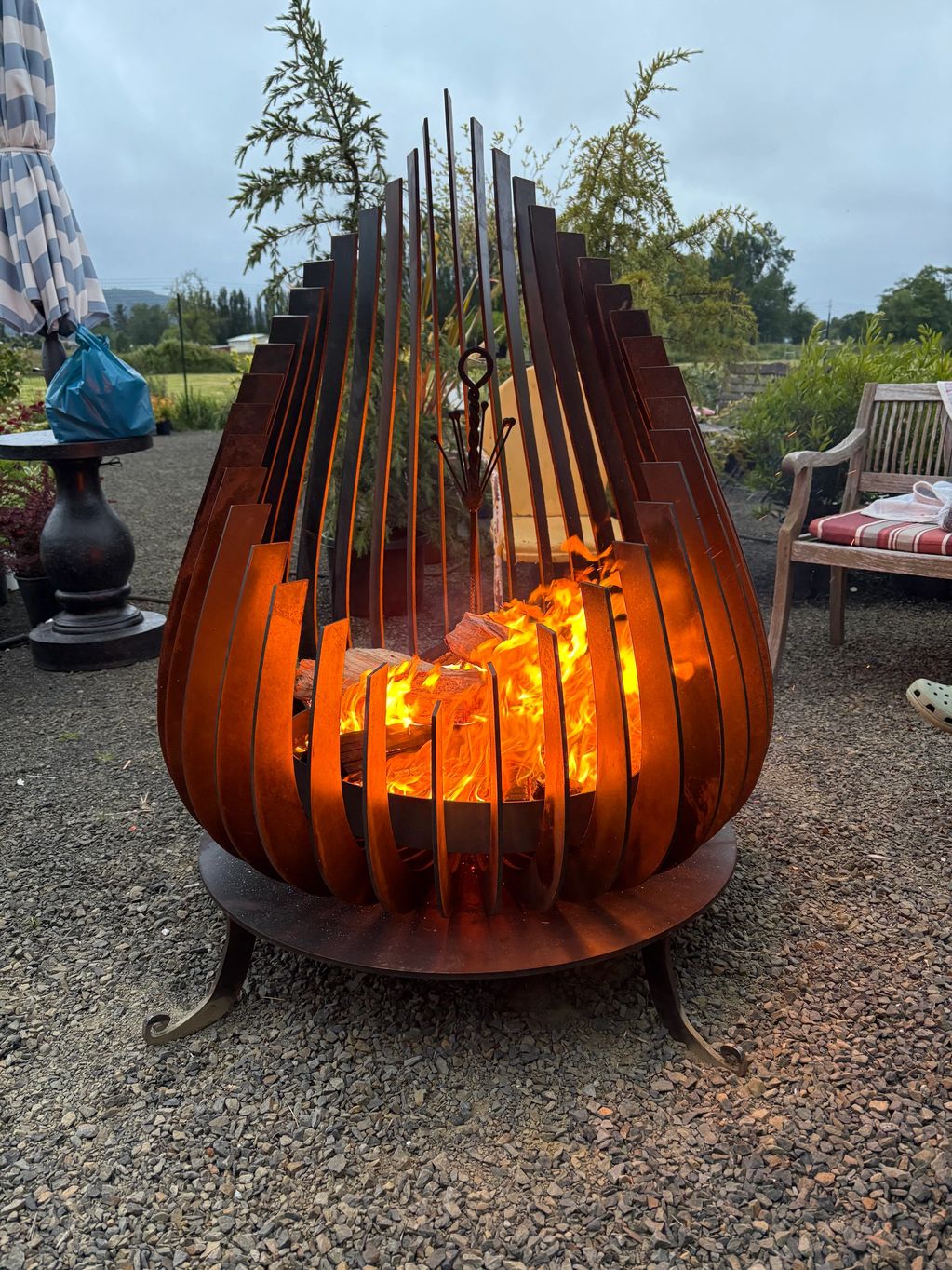 Modern metal fire pit with roaring flames on gravel patio.